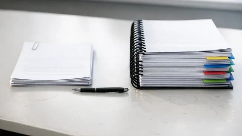 Two stacks of business documents on a meeting table - one thin stack representing Cyber Essentials assessment and one thick tabbed stack representing Cyber 365 programme materials